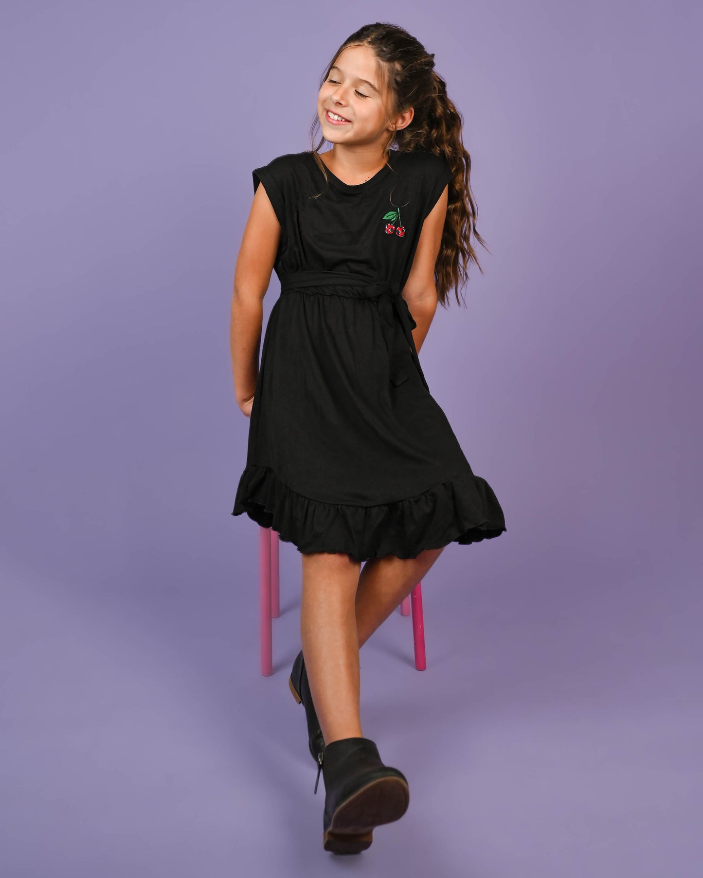 A happy young girl twirling in a black Cherry Skull Dress with charming ruffle details and waist tie, on a purple backdrop.