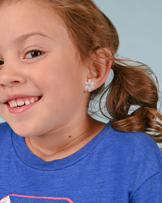 A young girl smiling, wearing winter-themed snowflake stud earrings, adding a festive touch to her casual blue tee.