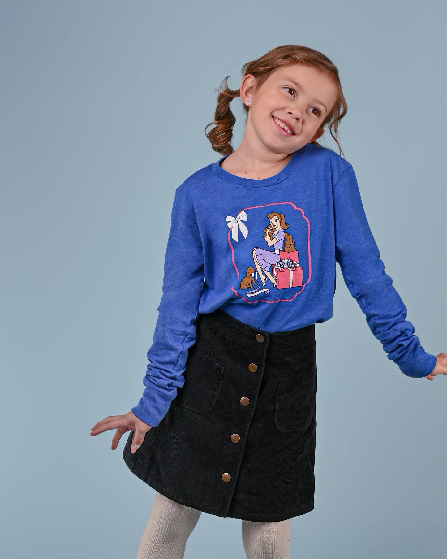 Cheerful young girl posing in a bright blue 'Puppy Love' long-sleeved tee, featuring a vintage print of a girl with her puppies, against a light blue background.