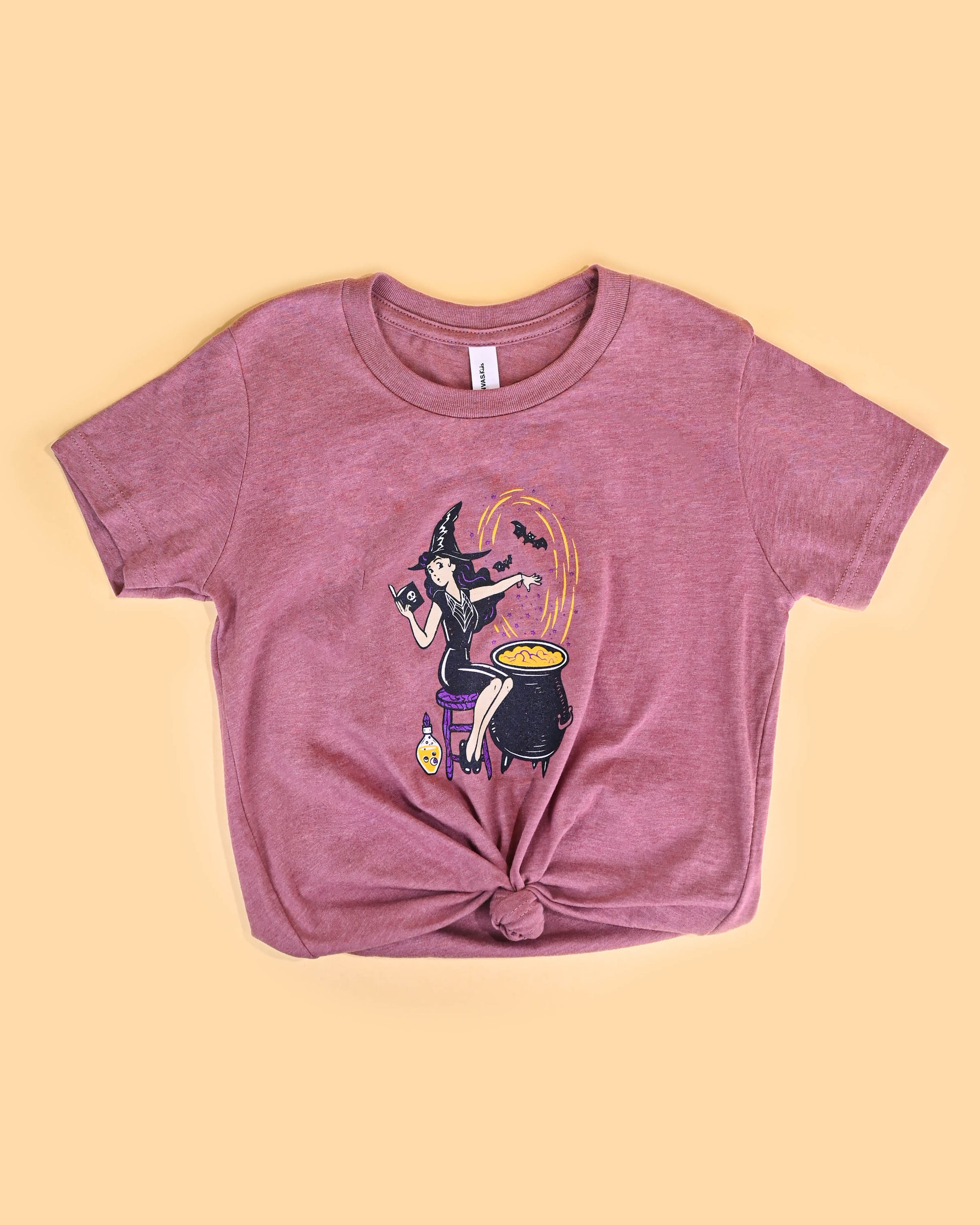 Mauve 'The Potion Maker' T-shirt with a playful witch graphic, neatly folded on a yellow background.