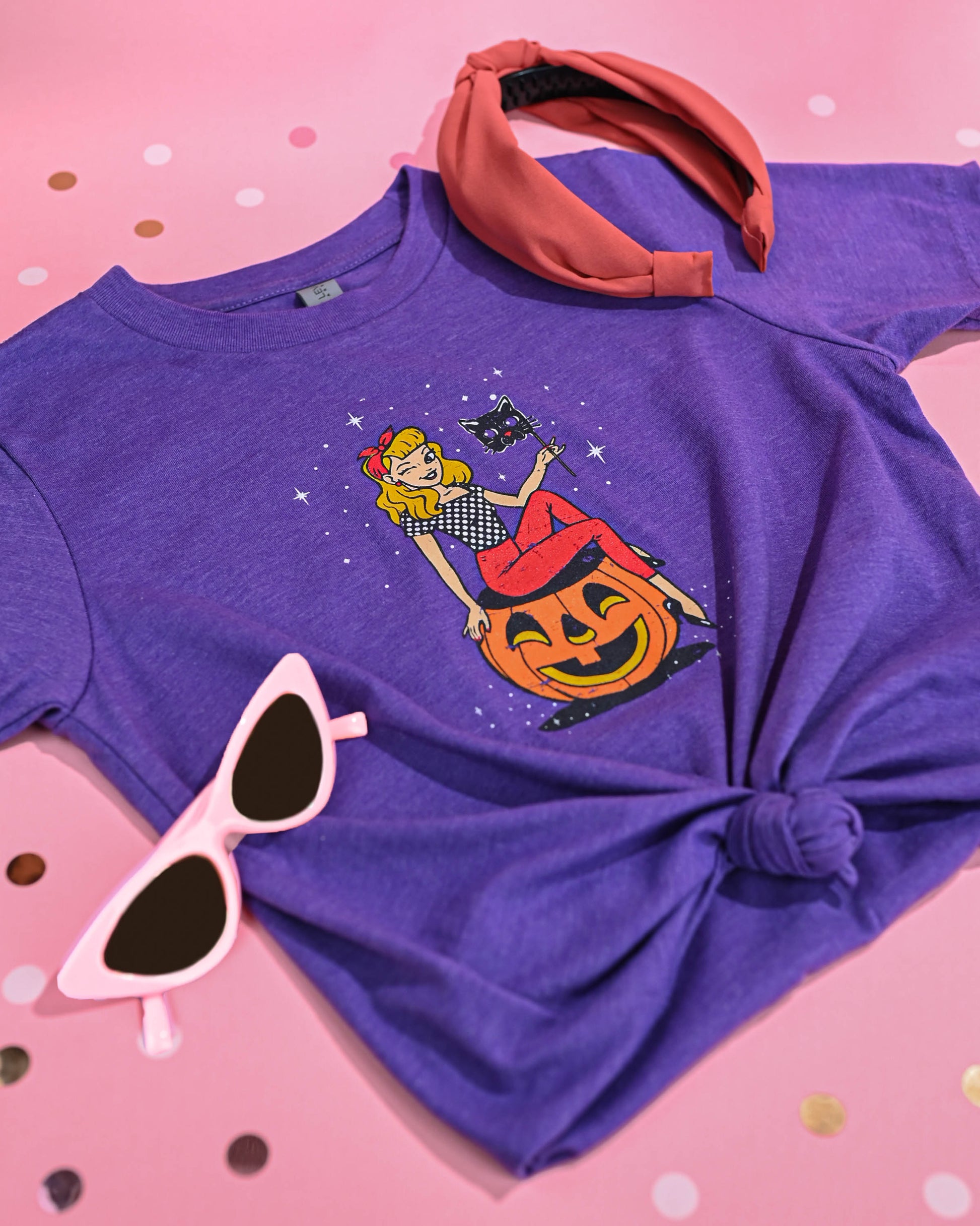 Stylized flat lay of the Black Cat Masquerade T-Shirt in purple, accessorized with a red headband and white sunglasses on a pink polka-dotted background.