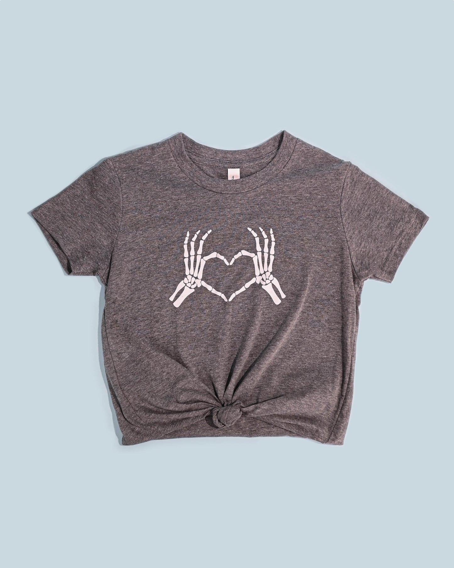 Alt text for image with a T-shirt: "Gray Skeleton Love Hands T-shirt laid flat on a blue background with a playful knot on the bottom.