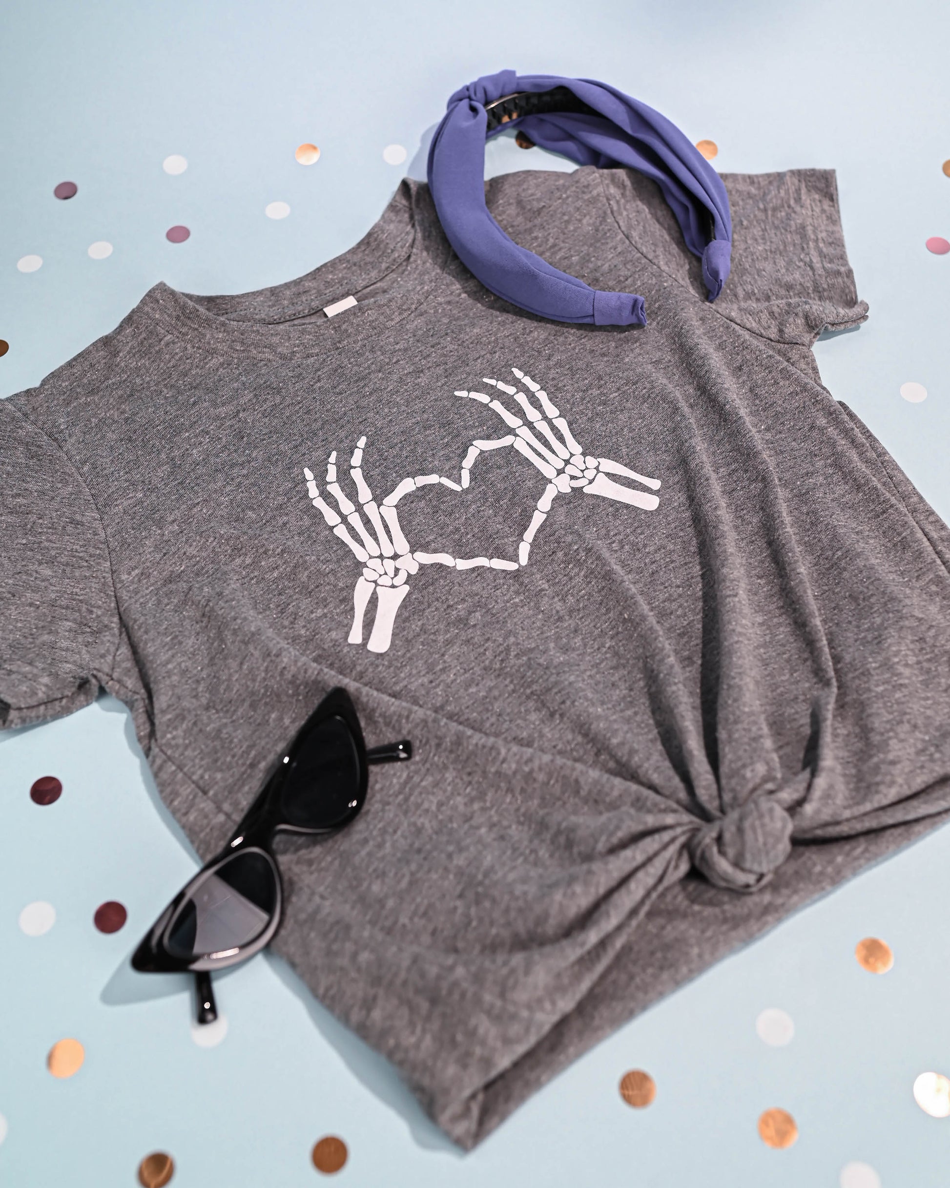 Alt text for styled image: "Skeleton Love Hands T-shirt styled with sunglasses and a blue headband on a polka-dotted pink background.