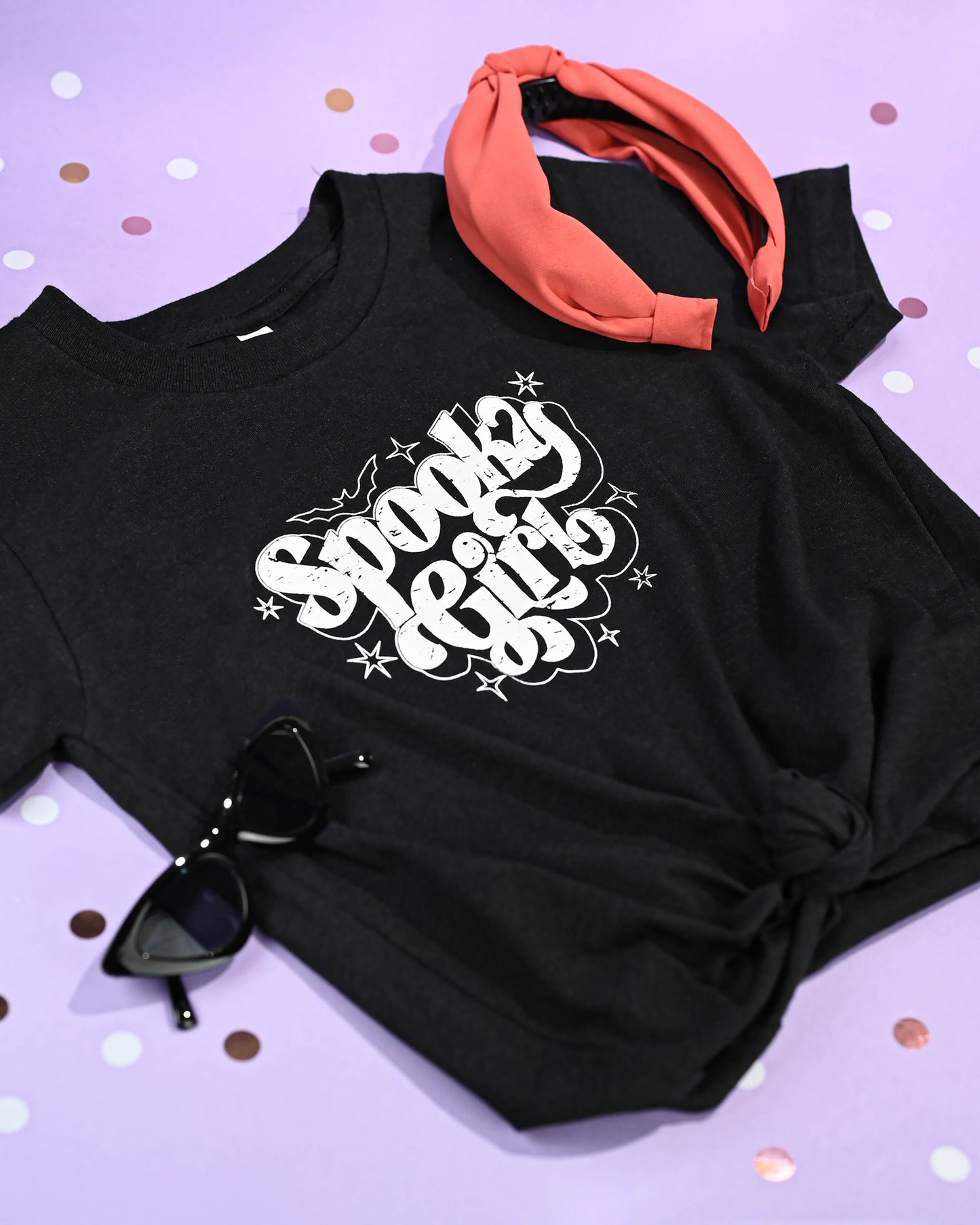 Black graphic tee with 'Spooky Girl' design displayed with pink sunglasses and orange headband on a dotted background.