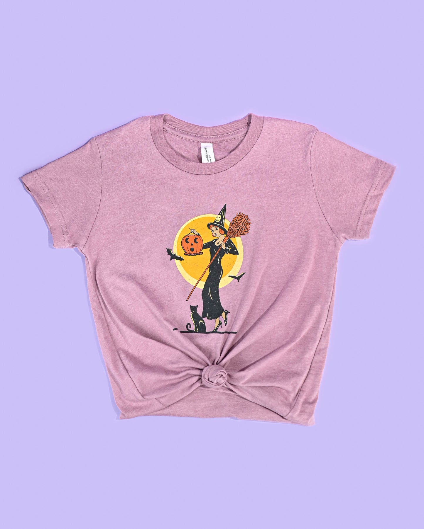 Soft mauve 'Witch with Jack-O-Lantern' T-shirt by Happy Donut with a playful Halloween graphic, knotted at the waist for a stylish look, on a purple background.