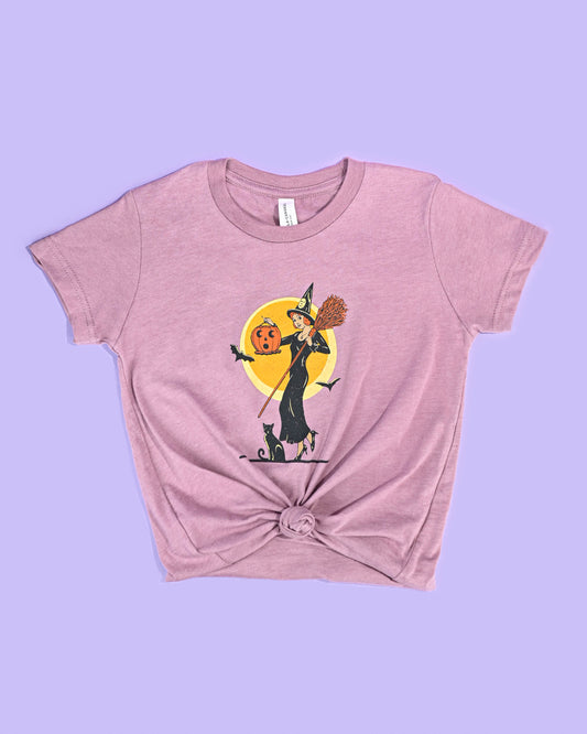 Soft mauve 'Witch with Jack-O-Lantern' T-shirt by Happy Donut with a playful Halloween graphic, knotted at the waist for a stylish look, on a purple background.