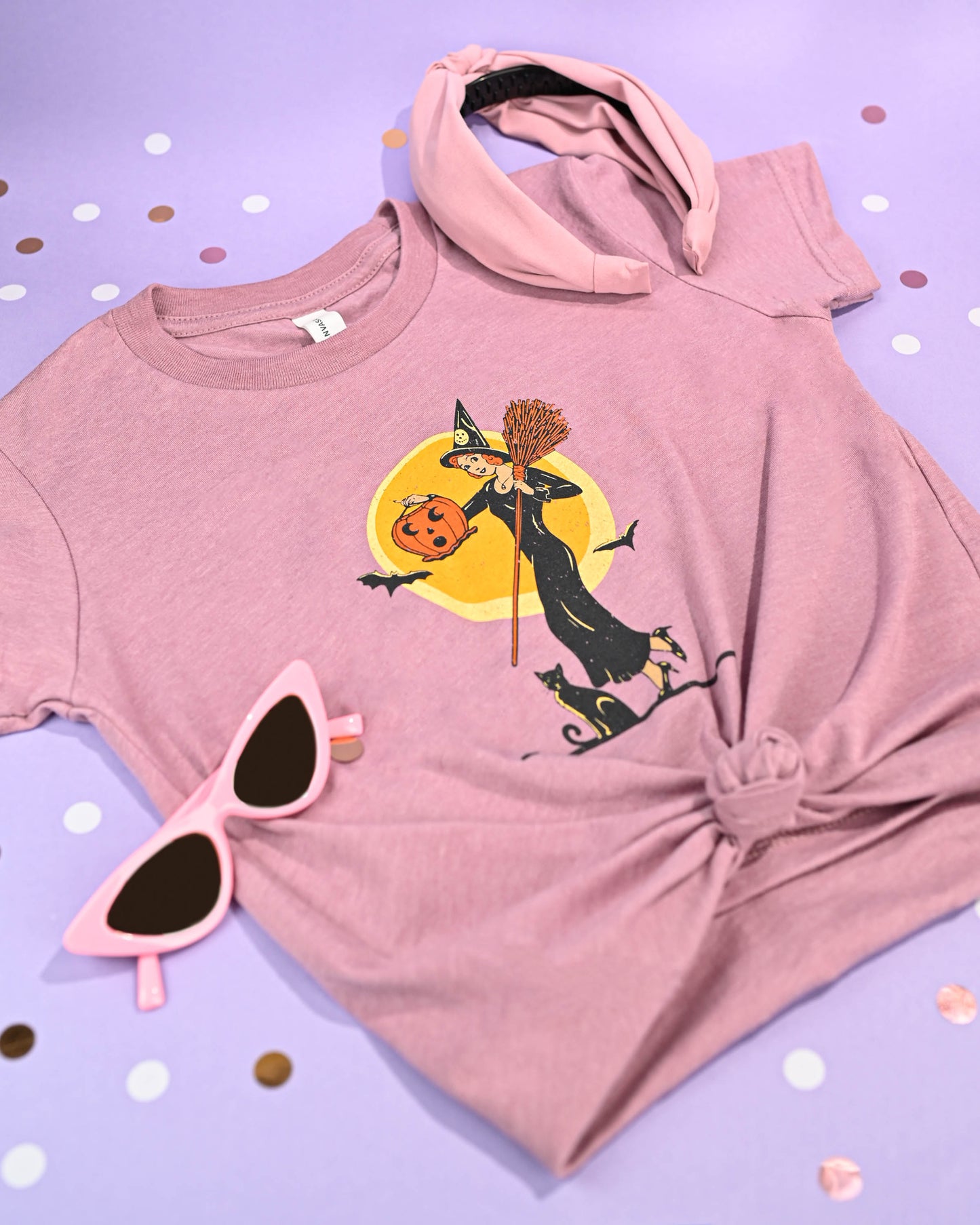 Happy Donut's 'Witch with Jack-O-Lantern' T-shirt displayed with a matching mauve headband and heart-shaped pink sunglasses, ready for a fun Halloween season.