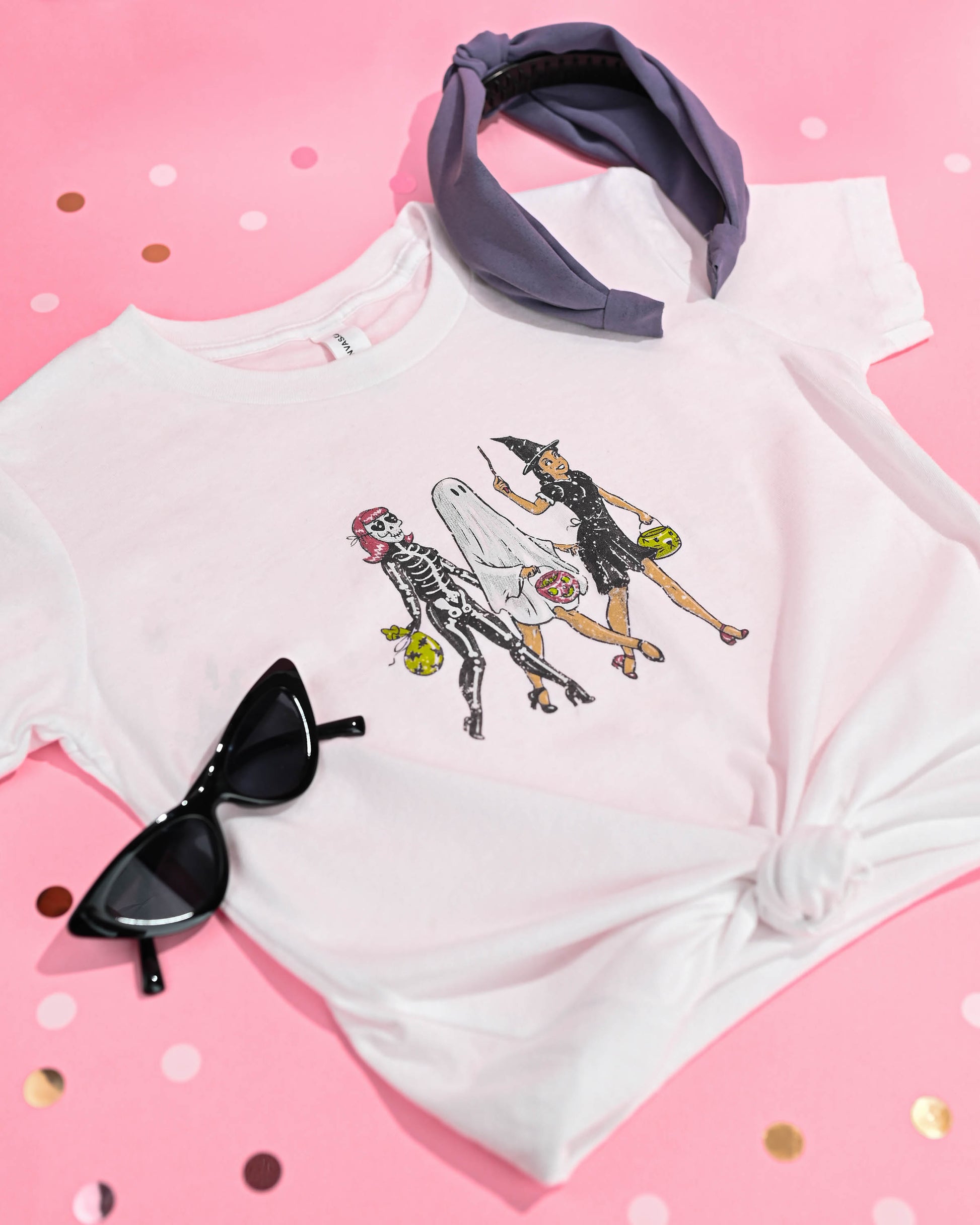 Stylish Happy Donut Trick or Treaters T-shirt laid flat with a purple headband and black sunglasses, showcasing the fun Halloween spirit on a pink polka dot background.