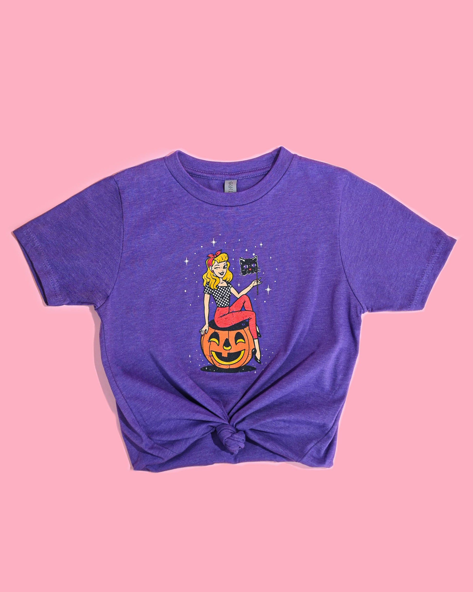 Black Cat Masquerade Tee in purple, featuring a knot style at the bottom, laid flat on a pink surface.