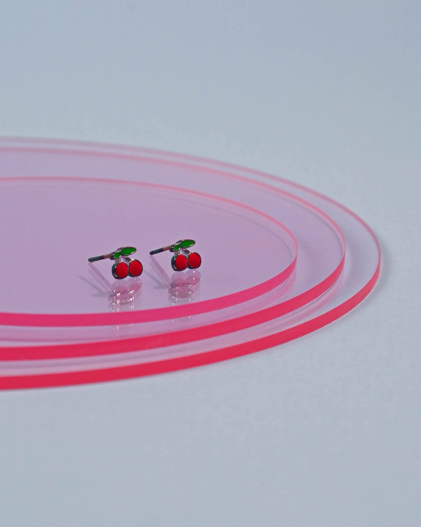 Vibrant cherry stud earrings with gleaming red enamel and silver stems, presented on a pink transparent concentric circle display.