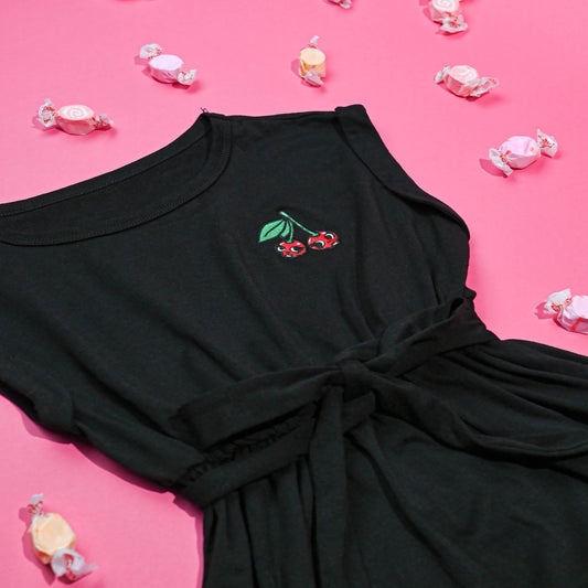 Black Cherry Tie Dress with a waist tie and ruffle detail, adorned with a cherry skull embroidery, surrounded by scattered candy wrappers on a pink background.