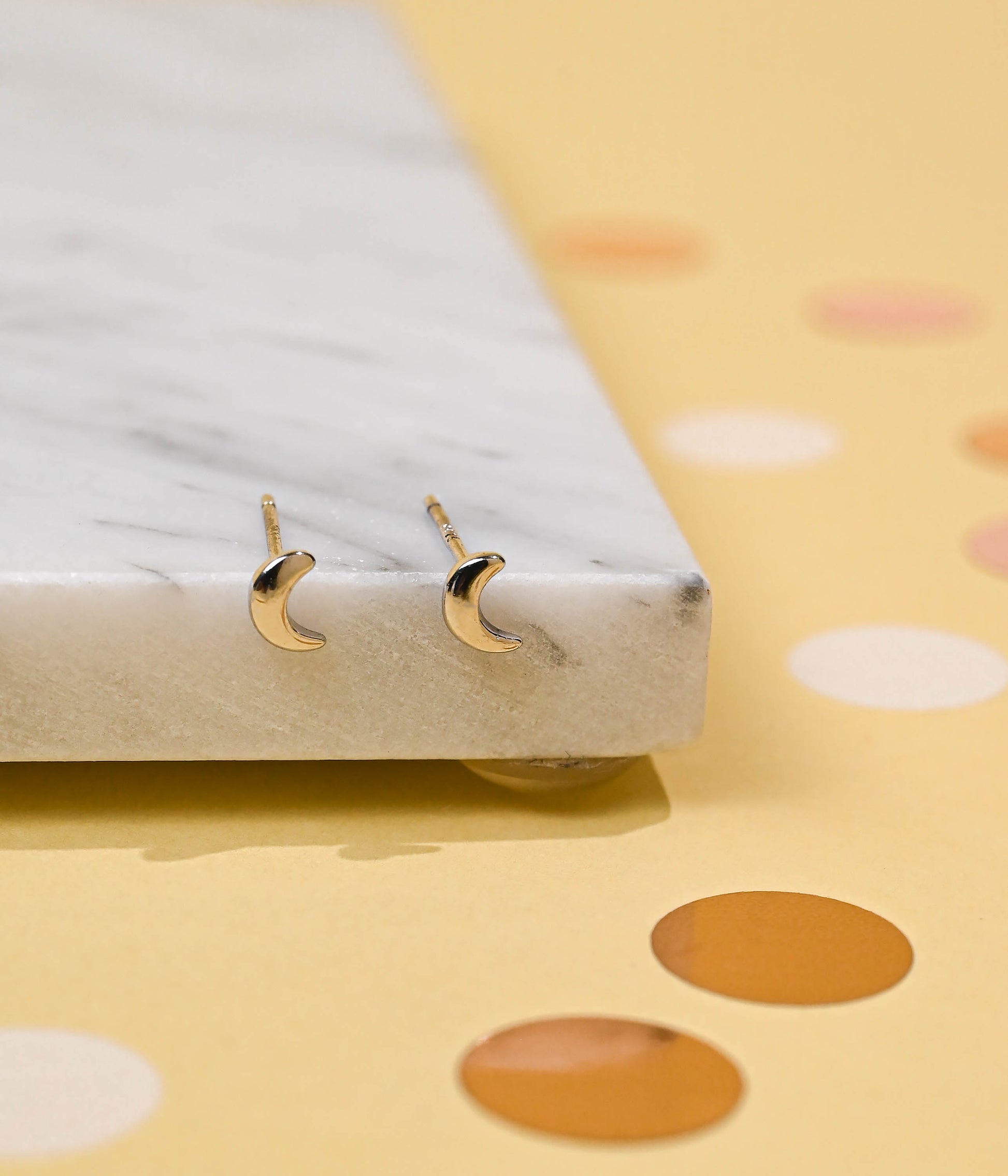 Elegant silver crescent moon stud earrings placed on a white marble surface with soft yellow background with pink dots.