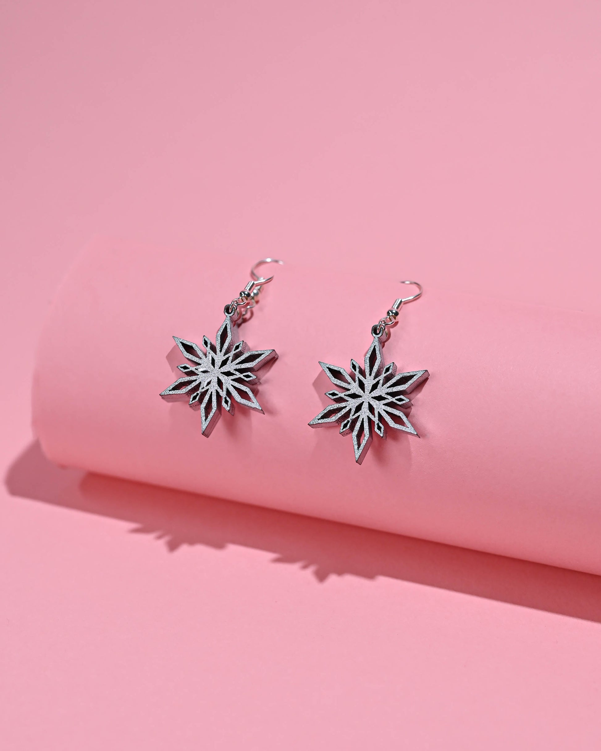 Silver snowflake dangle earrings displayed against a soft pink backdrop, emphasizing their shiny metallic finish.