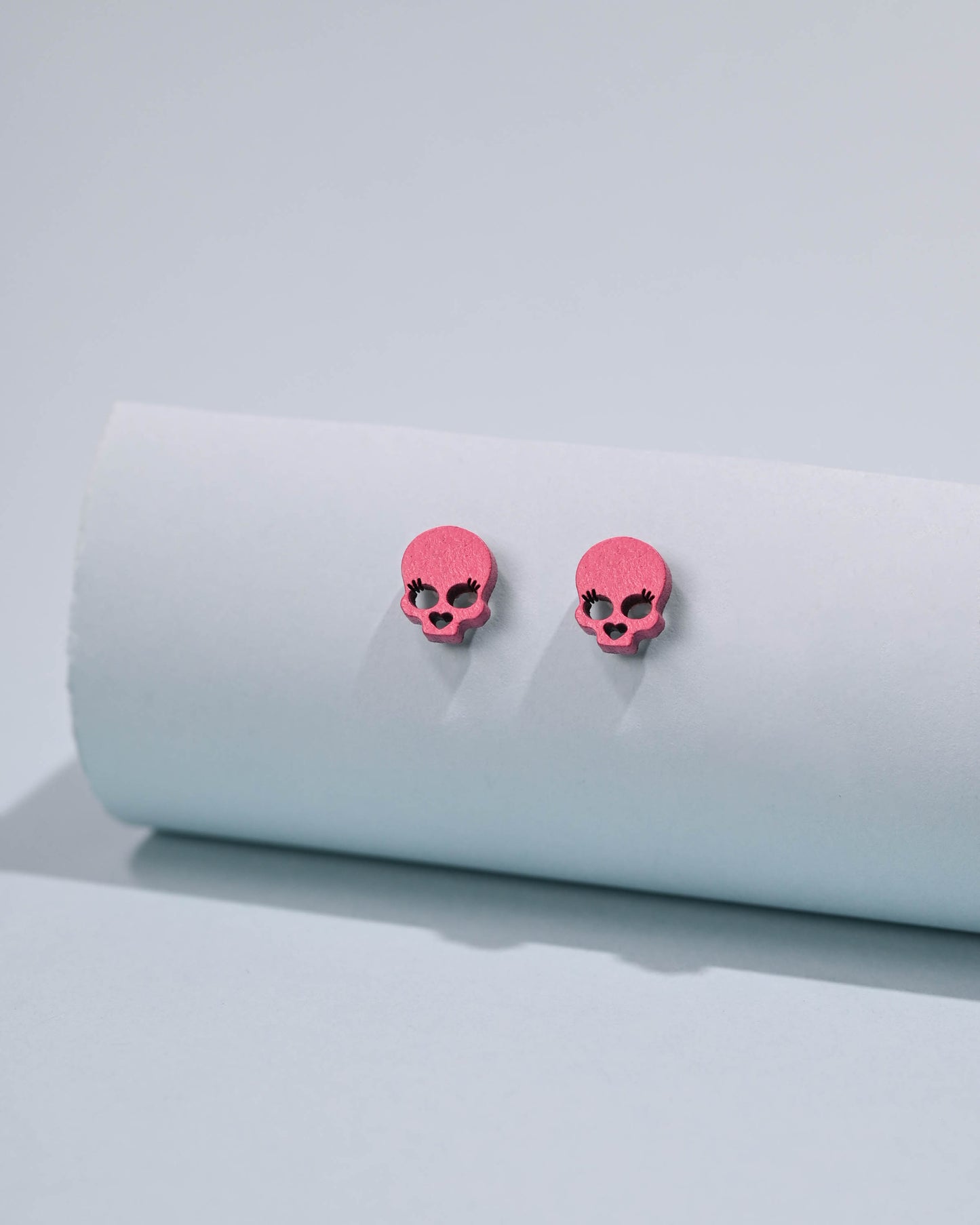 Pair of pink skull stud earrings resting on a white cylindrical surface, against a soft pink background, embodying a quirky yet charming style.