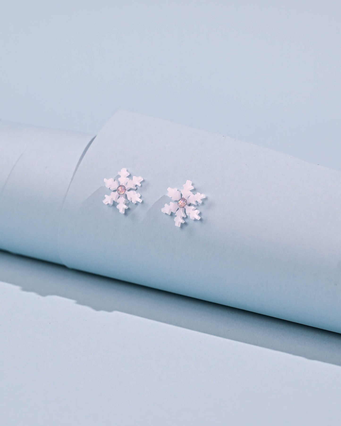 Close-up of snowflake stud earrings on a soft blue background, showcasing the white resin detail and stainless steel construction.