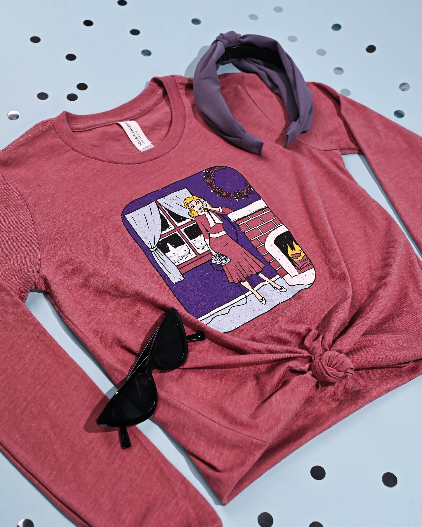 Stylish flat lay of a Gossip Girl long sleeve tee in mauve paired with a purple headband and black cat-eye sunglasses, on a polka-dotted pink surface.