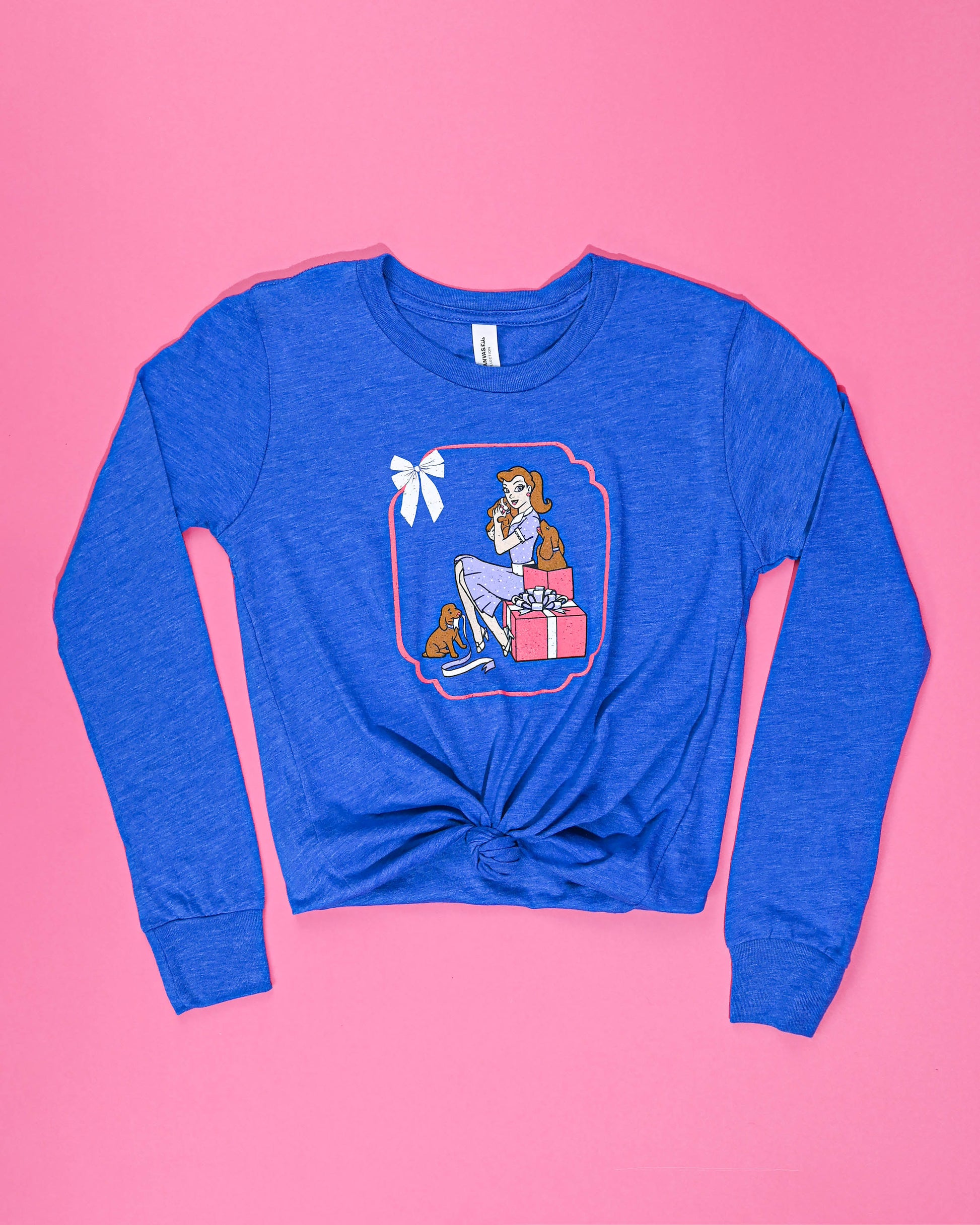 Blue long-sleeve 'Puppy Love' tee with a heartwarming vintage scene of a girl and her puppies, laid flat on a pink background.