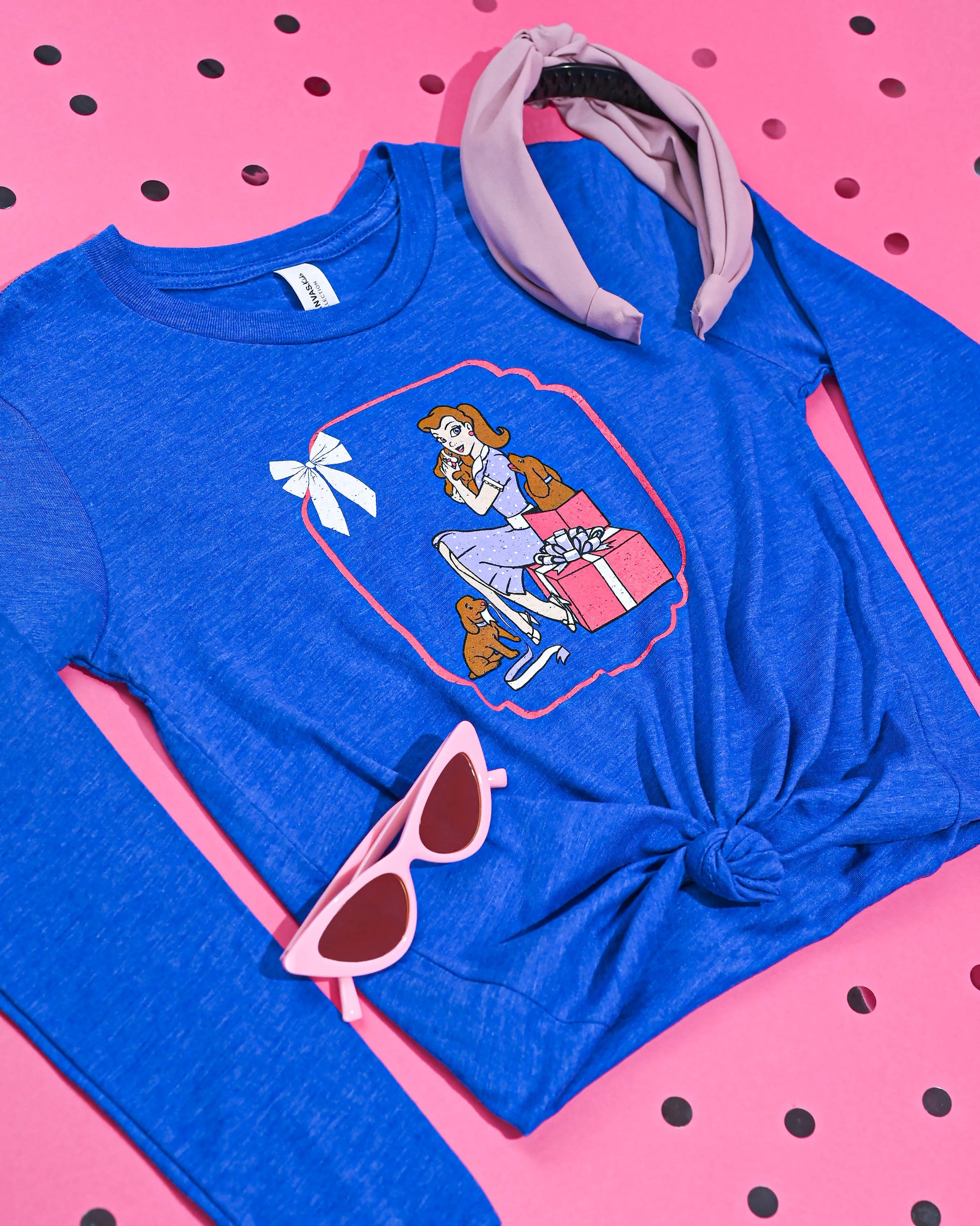 Stylish flat lay of the 'Puppy Love' long sleeve tee in blue, accessorized with a purple headband and pink cat-eye sunglasses, on a polka-dotted pink surface.