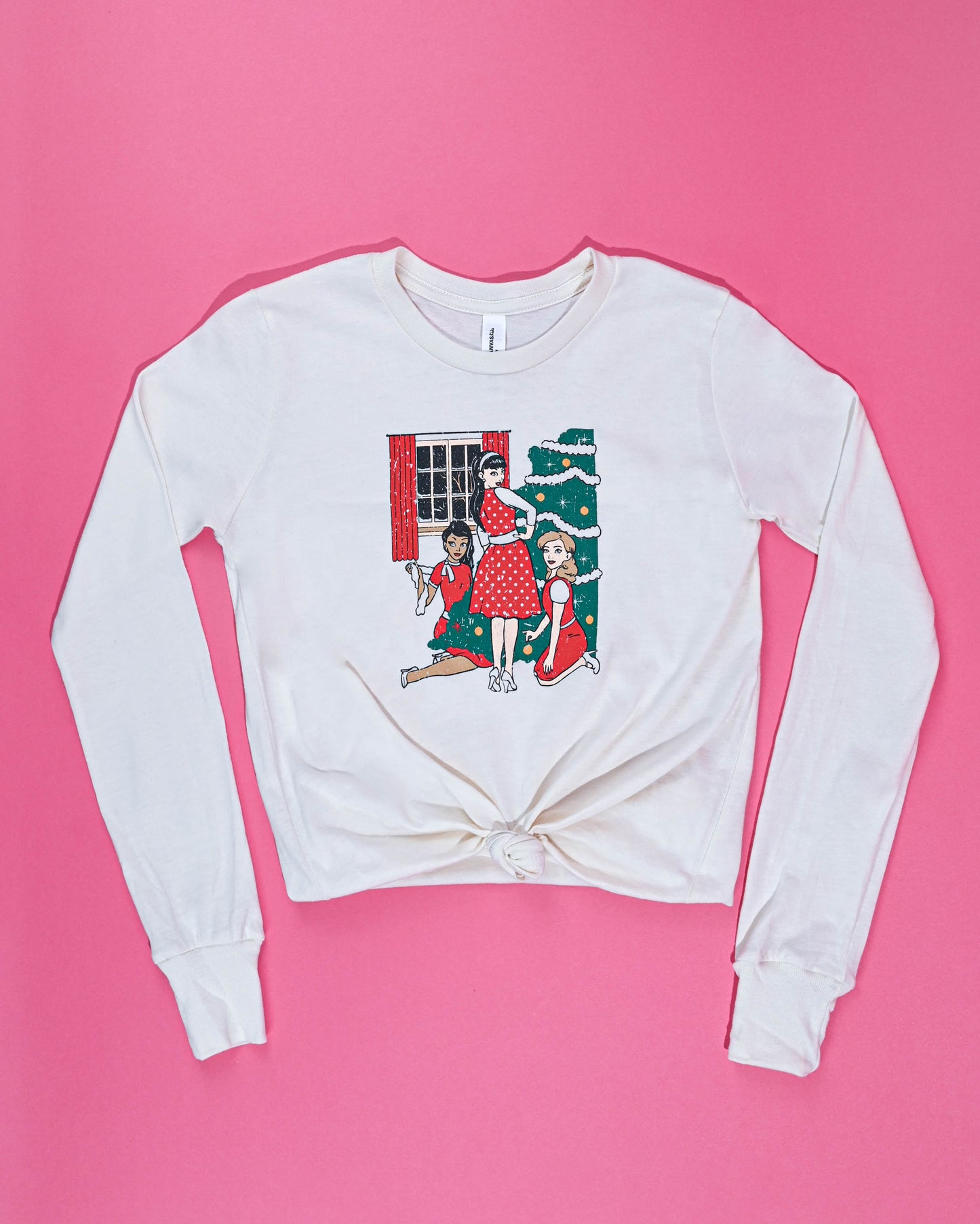 Festive long-sleeved white Christmas Trio Tee with a holiday tree graphic, laid flat against a vibrant pink background for a cheerful display.