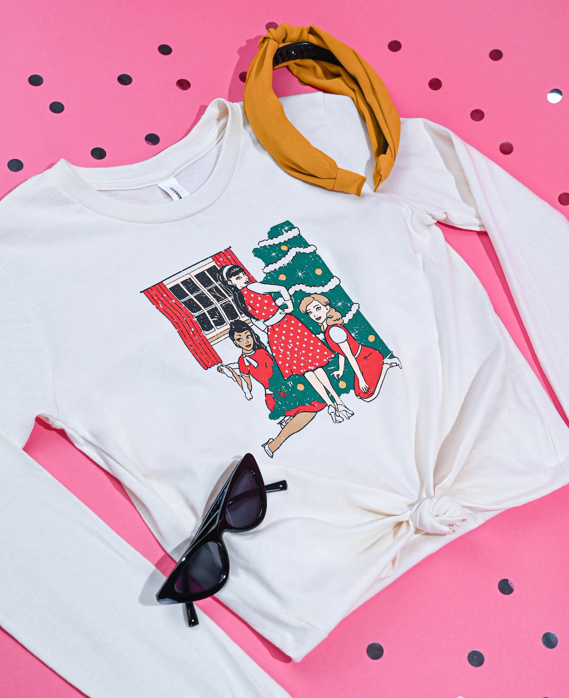 Vintage-inspired Christmas Trio Tee featuring three friends and a tree design, with a yellow headband and cat-eye sunglasses, on a pink polka dot surface.