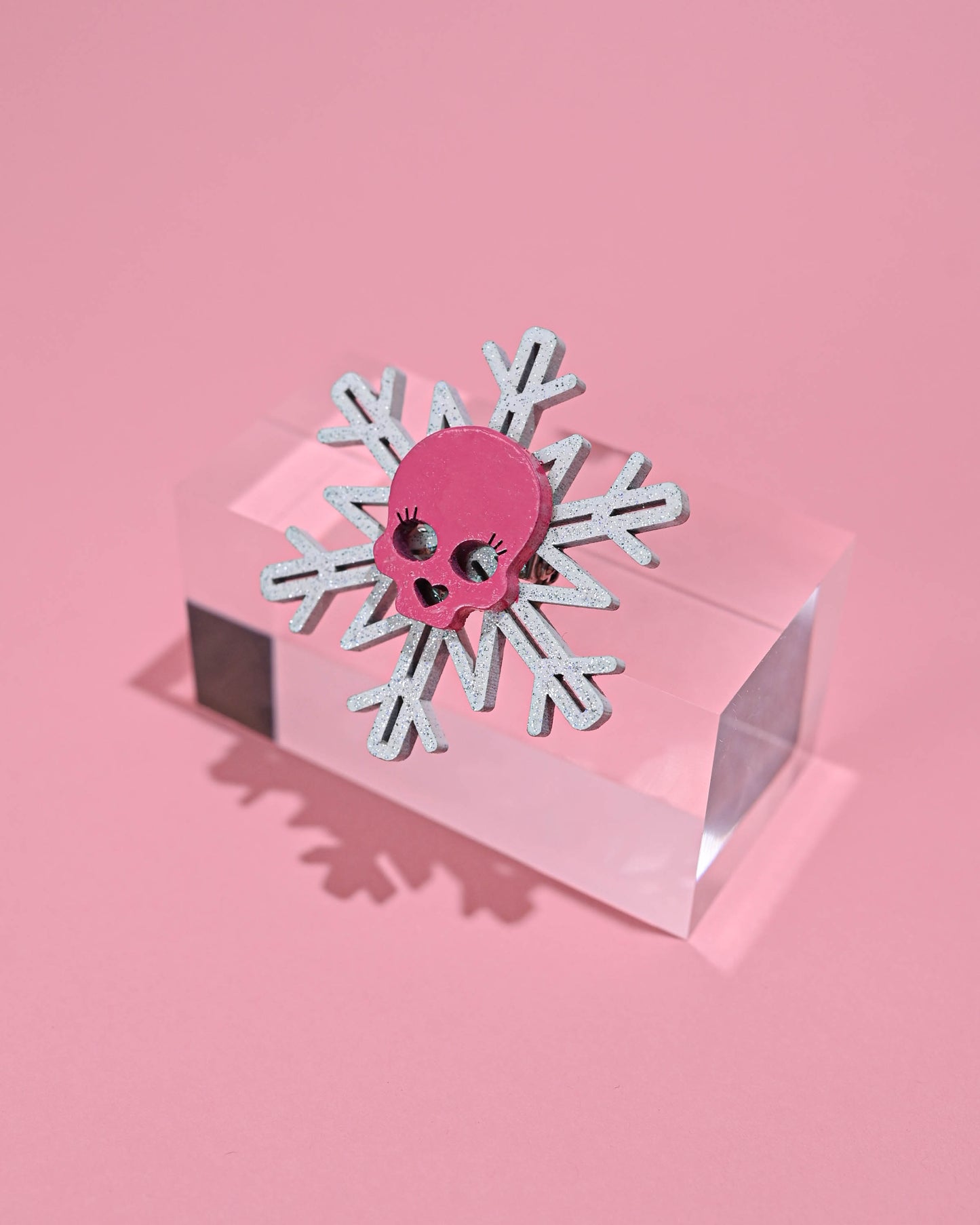 Pink skull snowflake pin with a sparkling touch, displayed on a geometric clear case against a soft pink background.