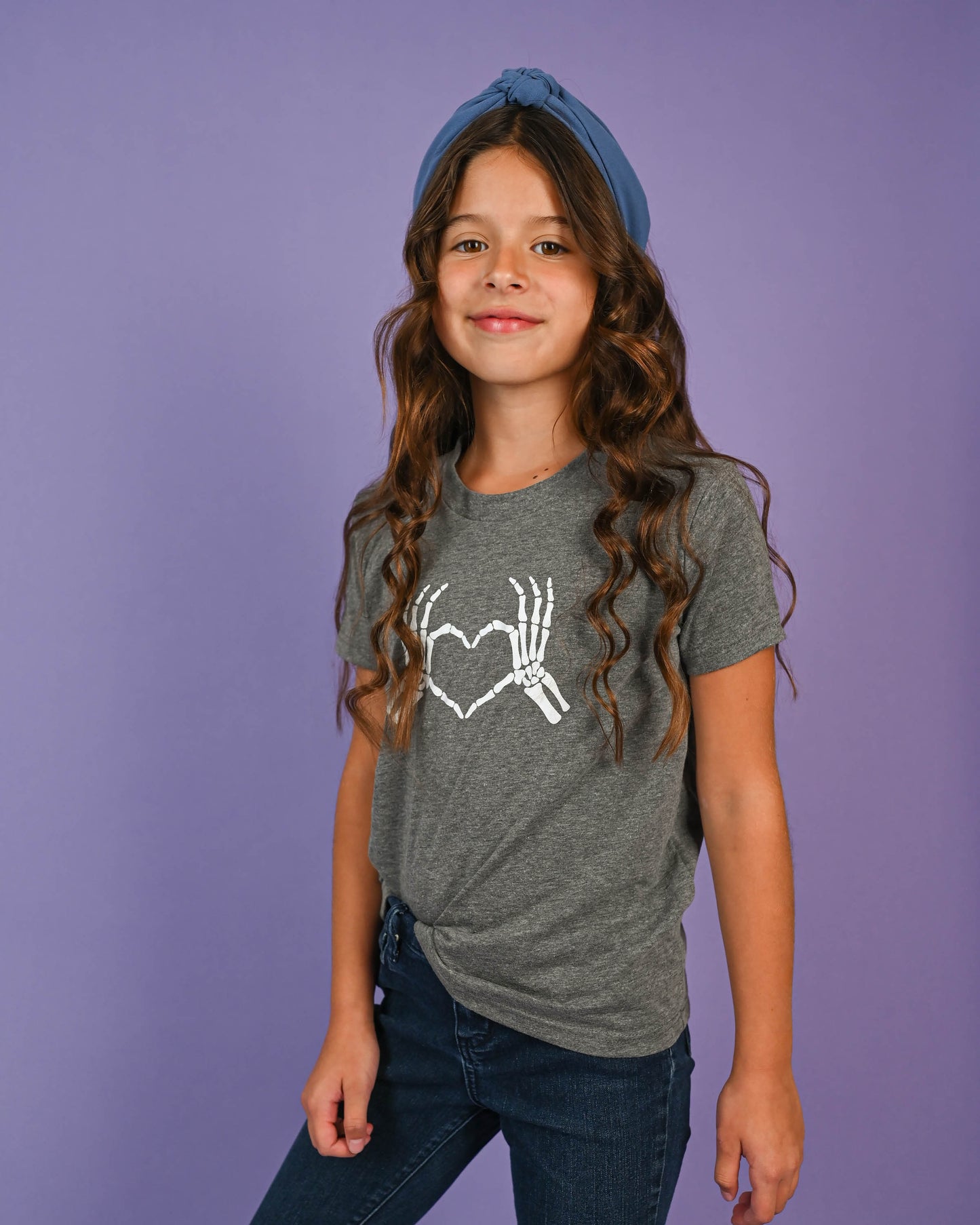 Young girl smiling, wearing the Skeleton Love Hands T-shirt, paired with a blue headband and jeans.