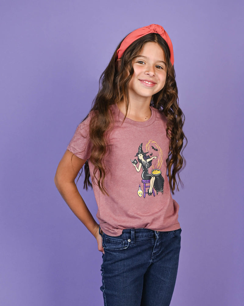 Smiling young girl posing in a mauve 'The Potion Maker' T-shirt with a witch and cauldron graphic, paired with jeans and a red headband. Perfect girls size 6-14 Halloween look.