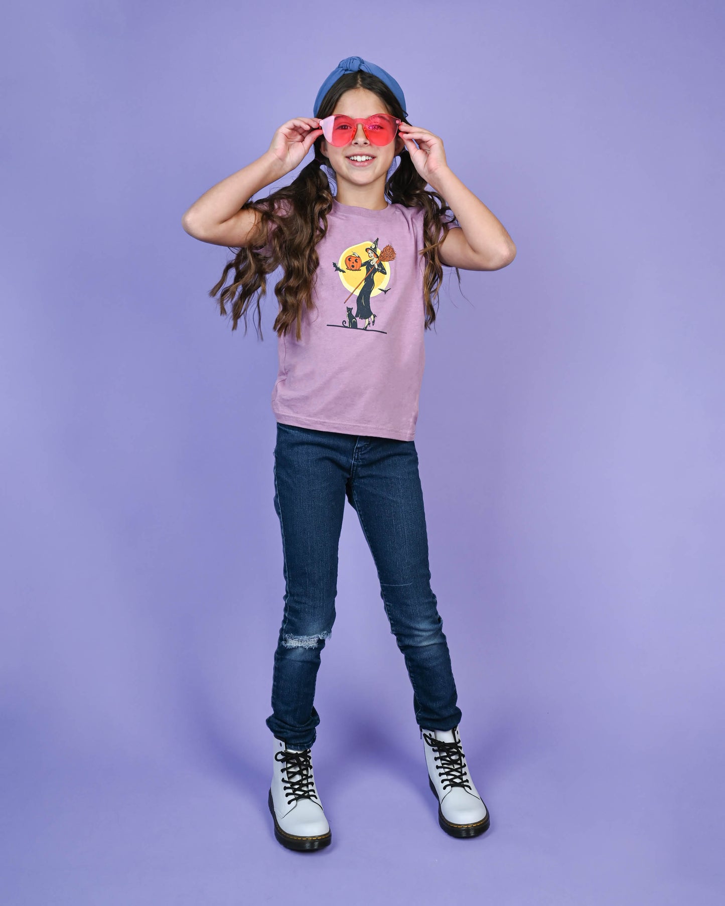 Young girl posing with a smile in Happy Donut's 'Witch with Jack-O-Lantern' T-shirt, complemented by her cool red sunglasses and casual jeans, embodying playful kids' fashion.