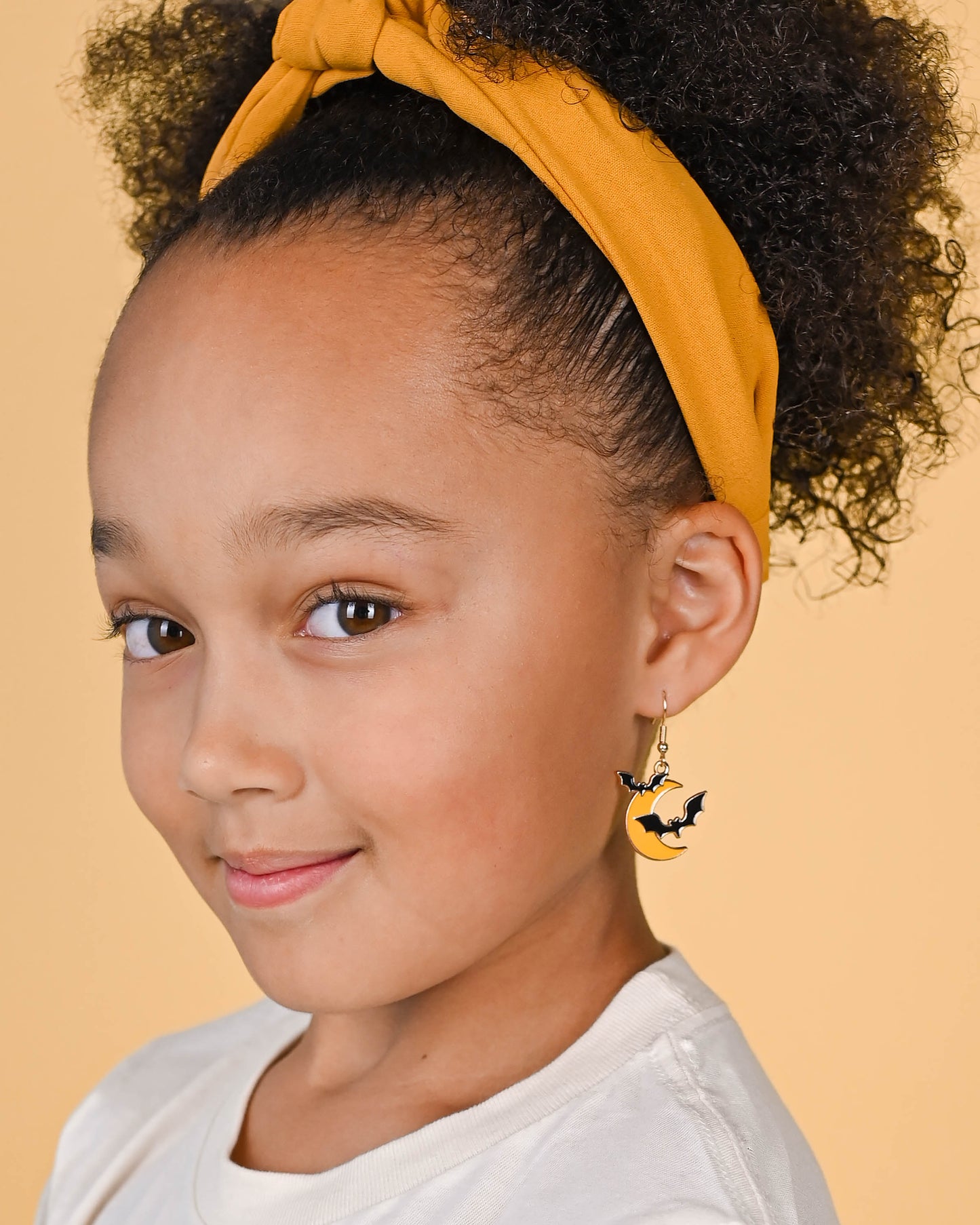 A smiling girl showcasing the Bats At Moonlight Dangle Earrings, paired with a yellow top knot headband and a white tee.