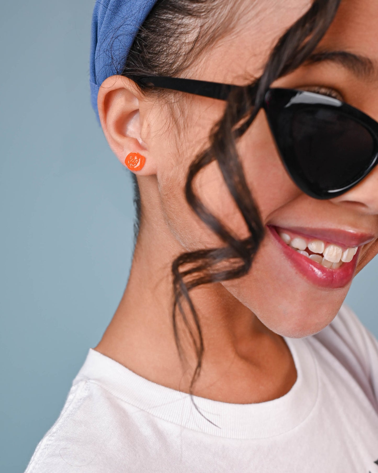 Girl wearing Jack-o'-lantern earrings, smiling playfully with cat-eye sunglasses and a blue headband, ready for Halloween fun.