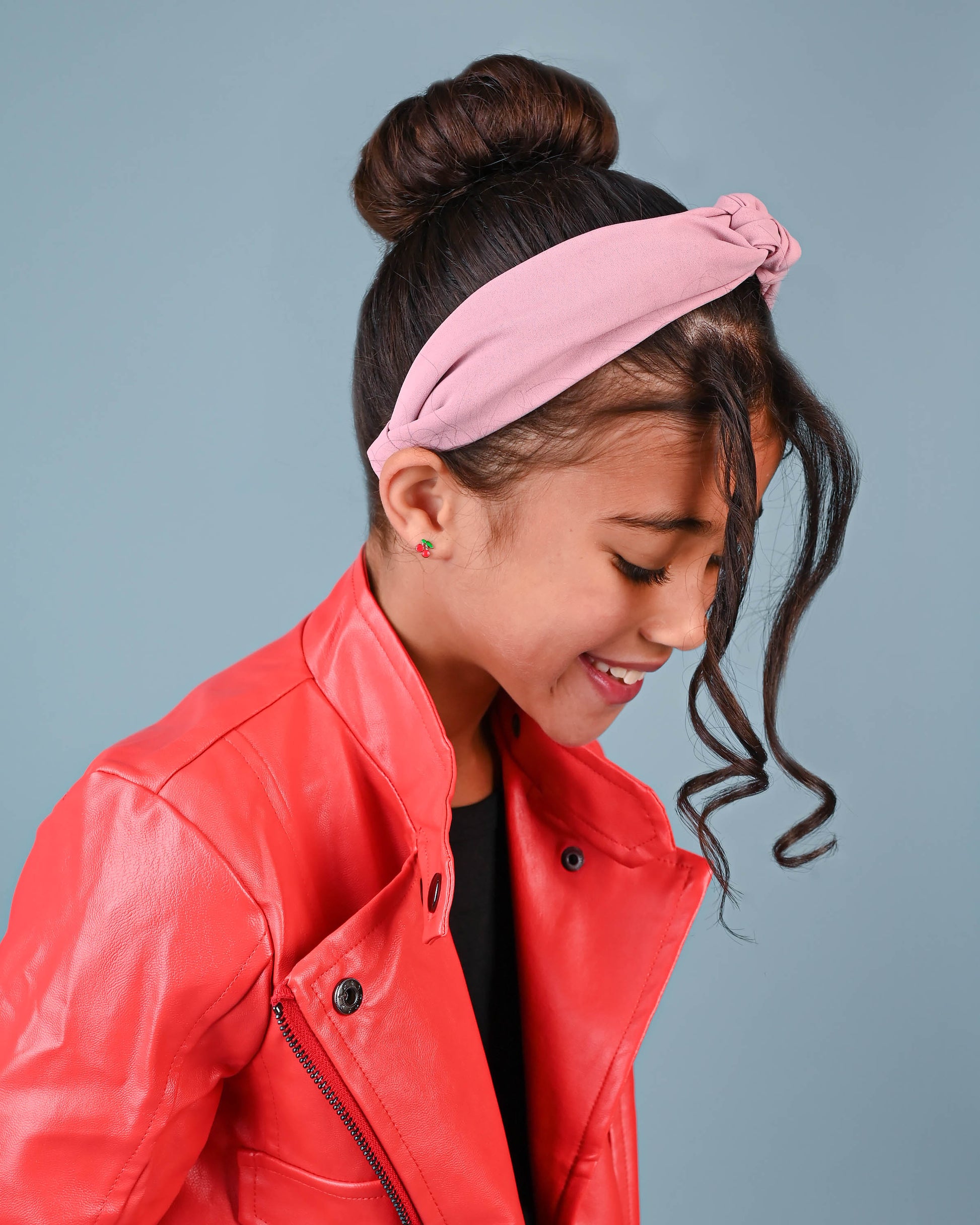 A joyful young girl with a pink headband smiles downward, showcasing her cherry stud earrings, adding a pop of color to her chic red leather jacket.