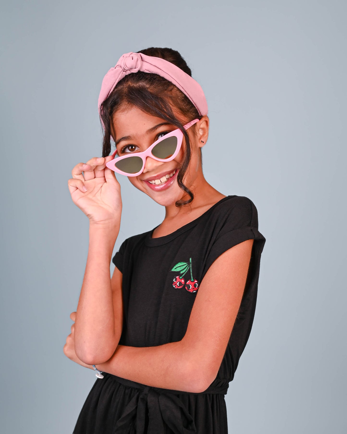 Playful girl tilting pink cat-eye sunglasses, wearing a pink headband and a black dress with a cherry design, ready for a sunny day.