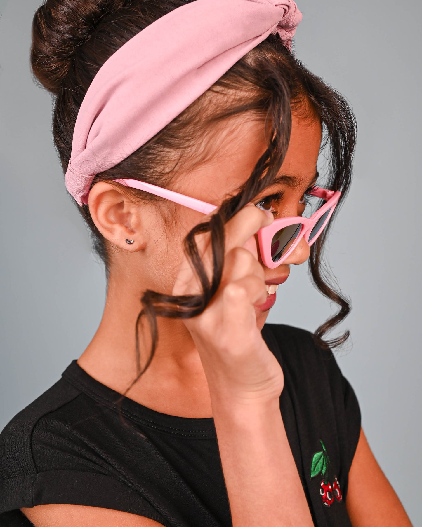 Young girl smiling sideways wearing pink headband and black top, with silver crescent moon earring visible on her left ear.