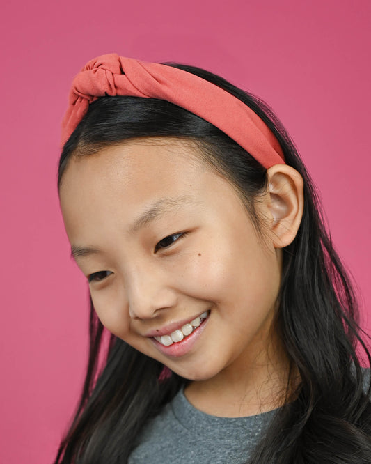 Young girl smiling wearing a coral top knot headband against a pink background.