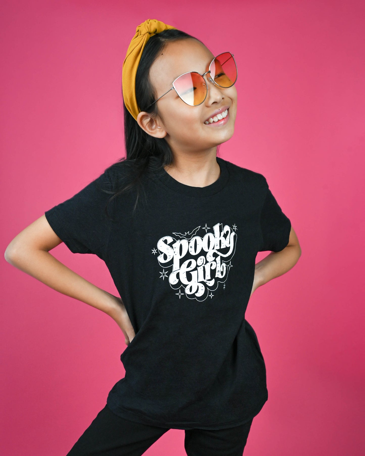 Young girl smiling wearing 'Spooky Girl' black t-shirt paired with yellow headband and reflective sunglasses.