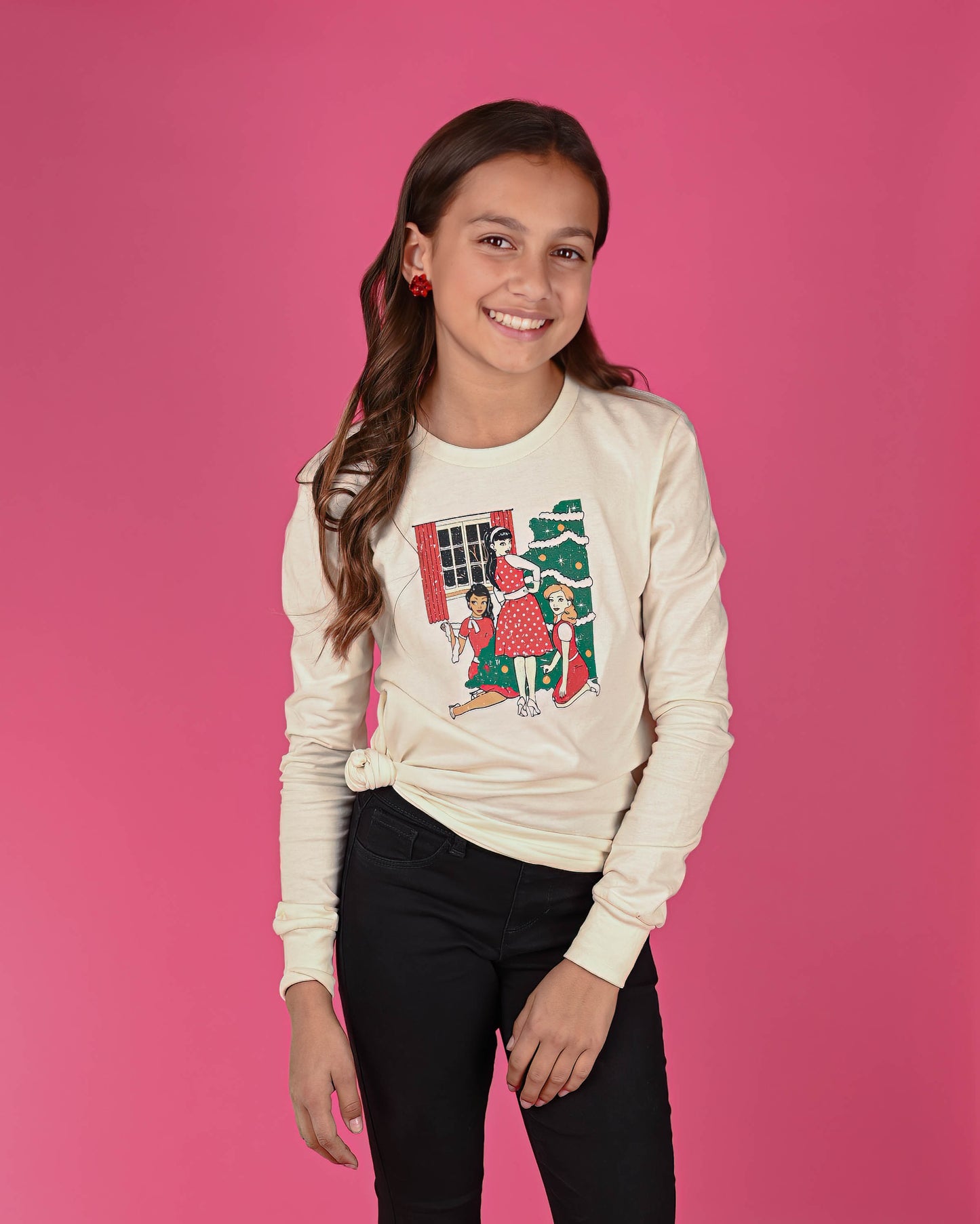 Smiling girl in a Christmas Trio long sleeve tee, paired with black pants and red bow earrings, against a pink background