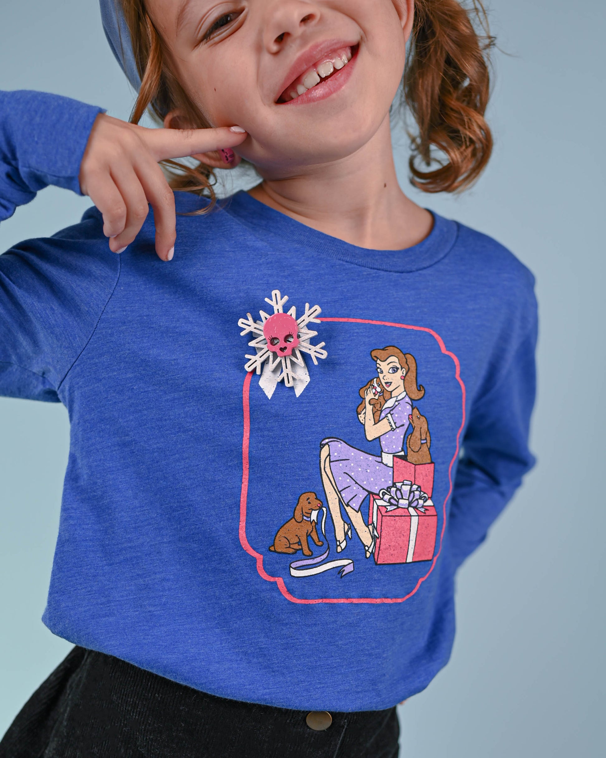 Cheerful young girl in a blue top smiling while pointing to her pink skull snowflake pin.