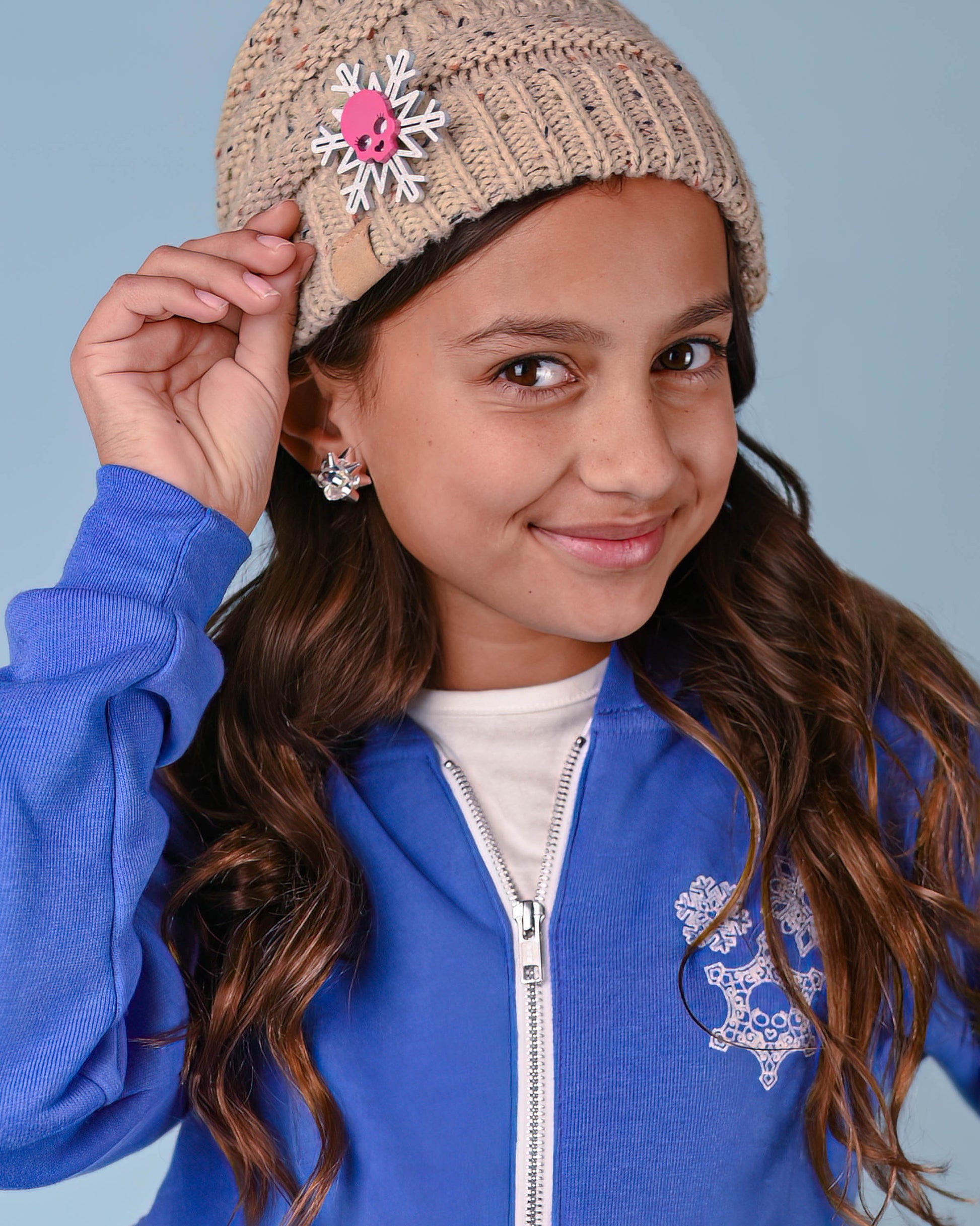 Close-up of a skull snowflake pin adorned on a beige knit hat worn by a young girl in a blue jacket.